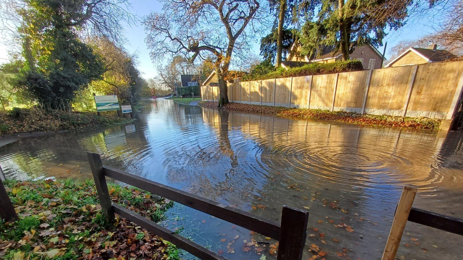 A flooded entrance to Harrold-Odell Country Park. There is a large amount of water on the road, trees surrounding the road, a large fence, wooden barriers and two signs for the park. The sky is blue and there are leaves on the ground. 