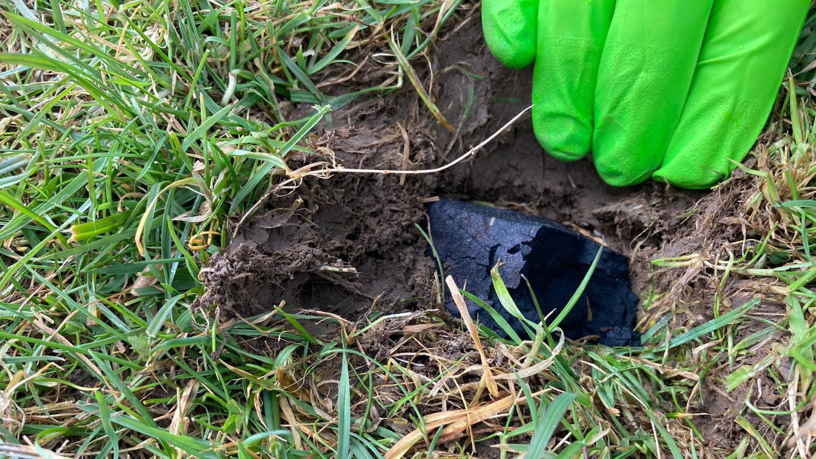 The Winchcombe meteorite fragment embedded in the ground in 2021. It is black and is partially buried under earth and grass. A hand covered with a bright green glove is holding back part of the earth to reveal the rock