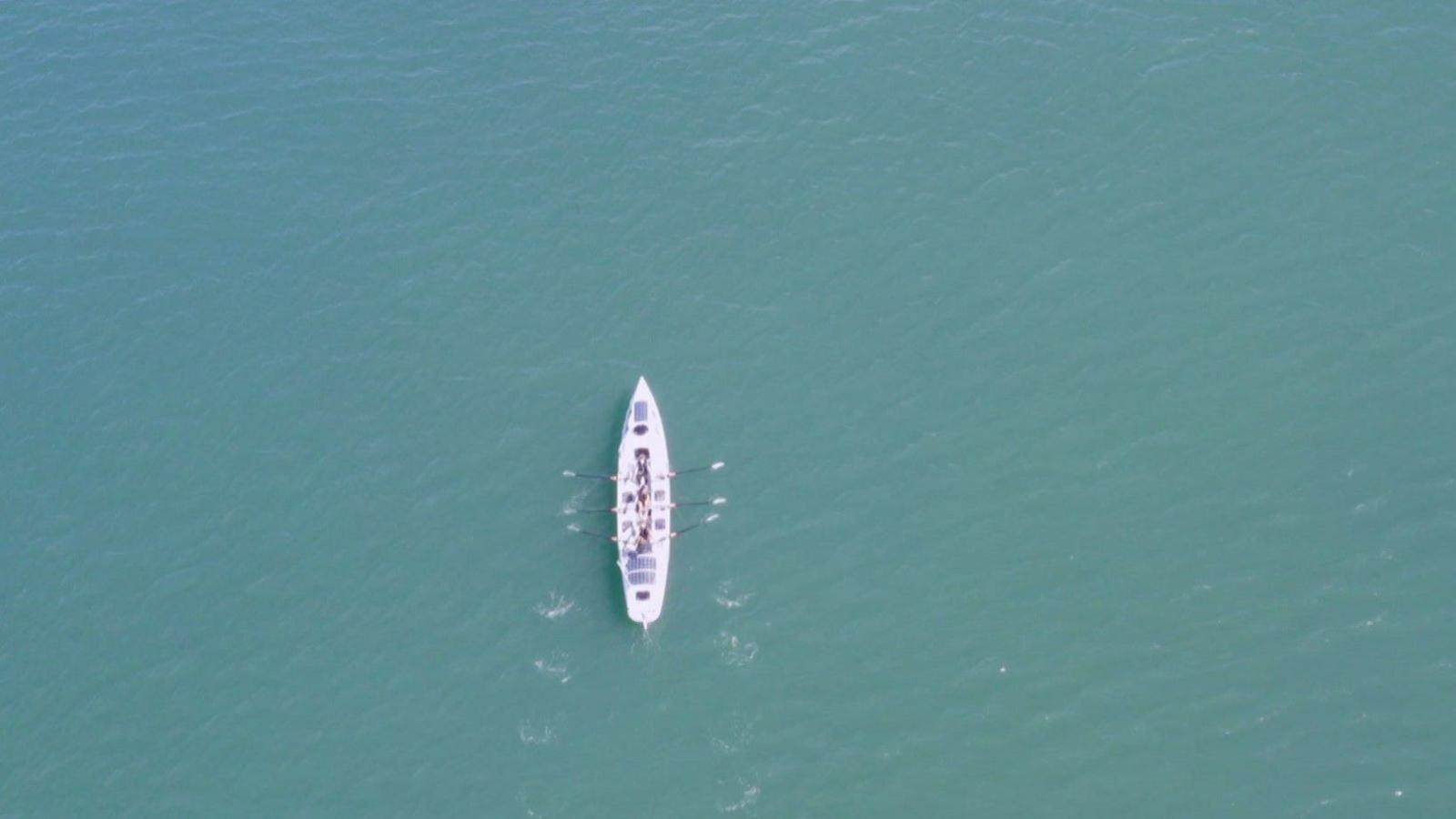 A rowing boat with four rowers in the middle of the ocean is viewed from the sky.