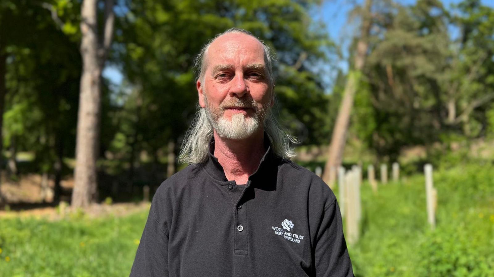A man with a grey beard and long grey hair tied back is wearing a black t shirt which says "Woodland Trust Northern Ireland" is stood in front of a woodland.
