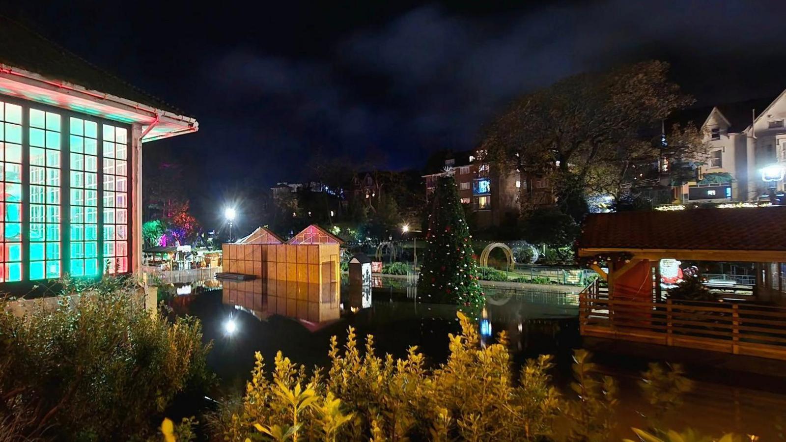 A flooded lower gardens taken at night. On a bank on the left is a rectangular glazed bandstand illuminated in blue. Structures and Christmas trees are in darkness and partly submerged.