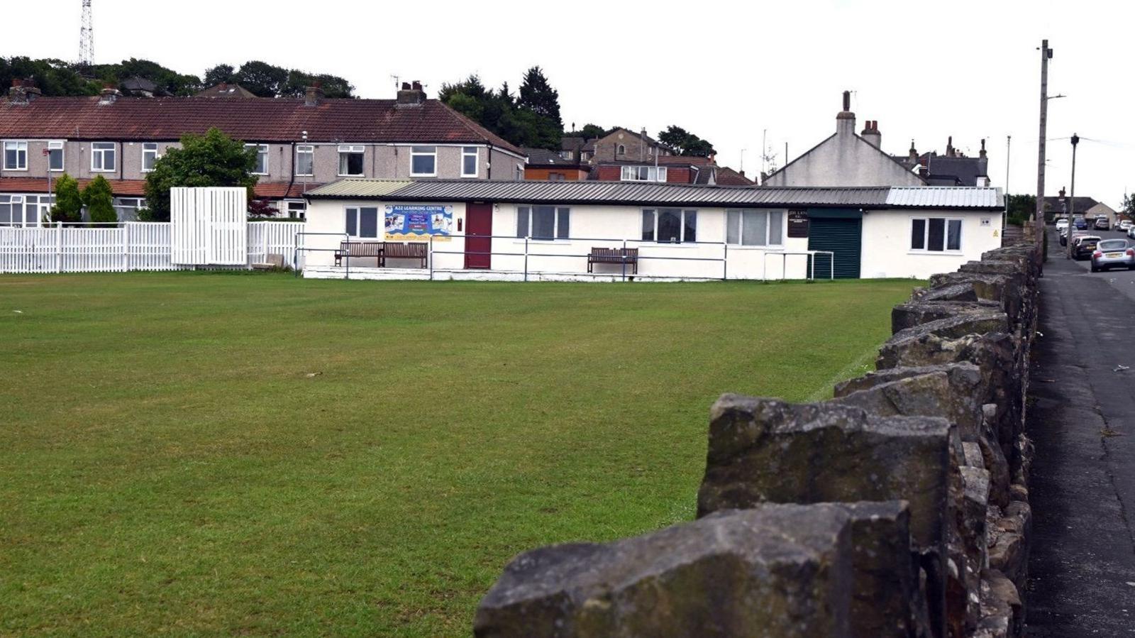 A single storey white building with a large green cricket pitch in front of it and a low stone wall on the right next to a road