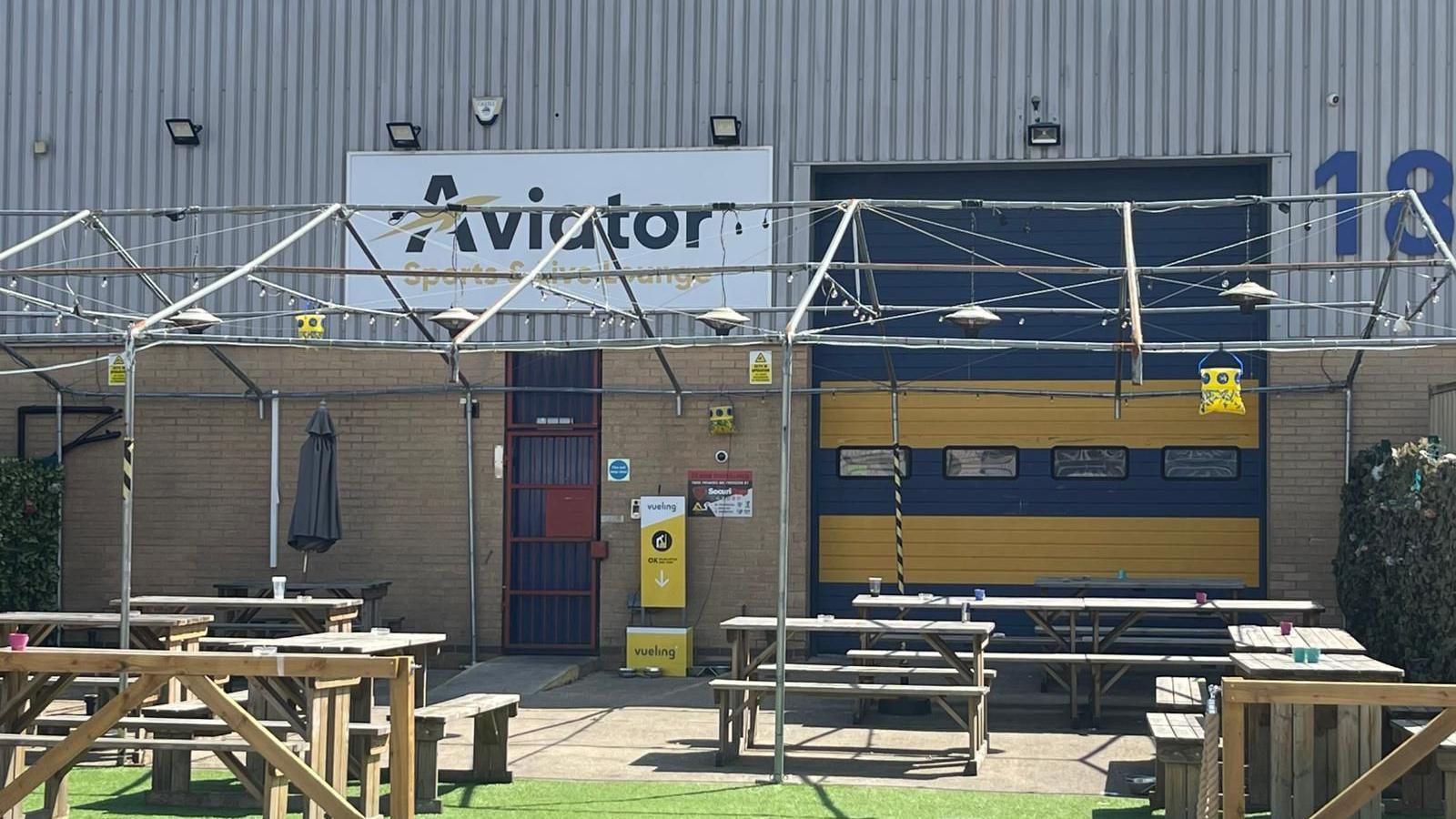 Aviator Sports and Live Lounge. A number of wooden benches are placed outside the entrance. The building looks like an industrial unit, with a lower half in brick and the top half clad in corrugated sheeting. It has a dak blue and yellow garage style roller shutter. To the left is a small door covered by a locked gate.