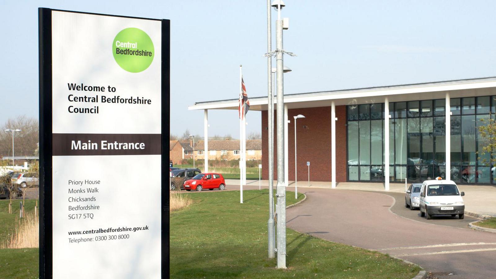 A picture of Central Bedfordshire Council Headquarters at Chicksands. The building, a modern glass-fronted construction, is in the background on the right hand side. On the left, in the foreground, is a sign saying "Welcome to Central Bedfordshire Council".