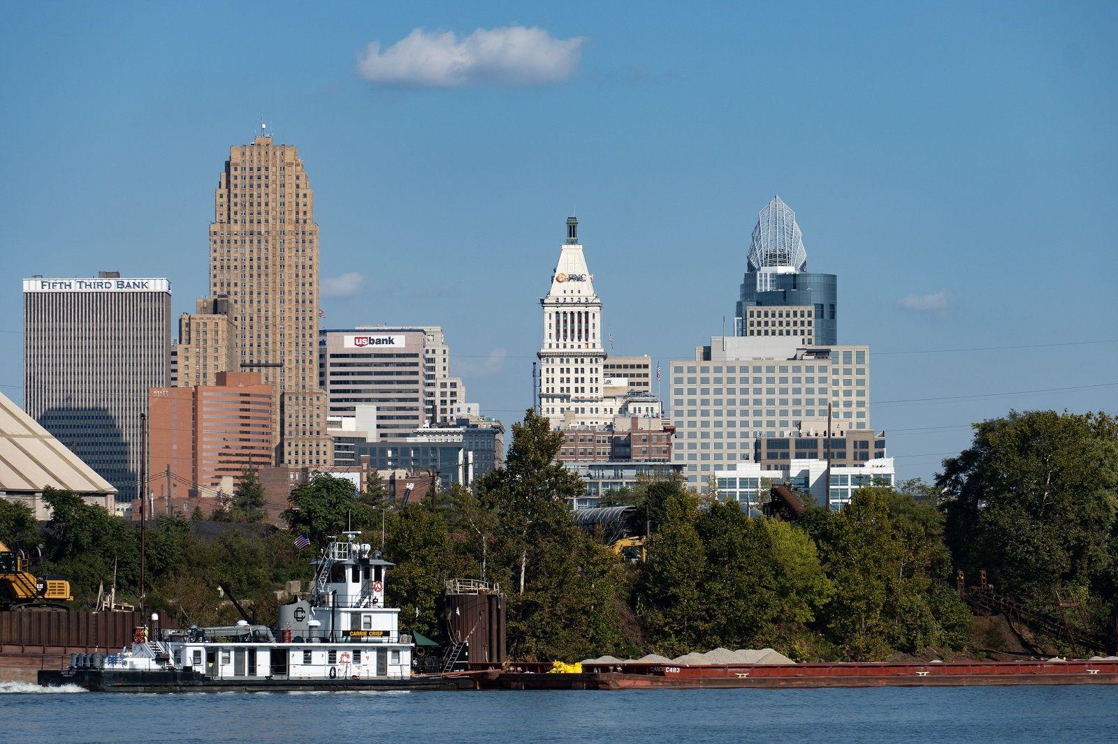 A white boat in the Ohio River in front of a cluster of trees and a skyline of several skyscraper buildings on a bright day