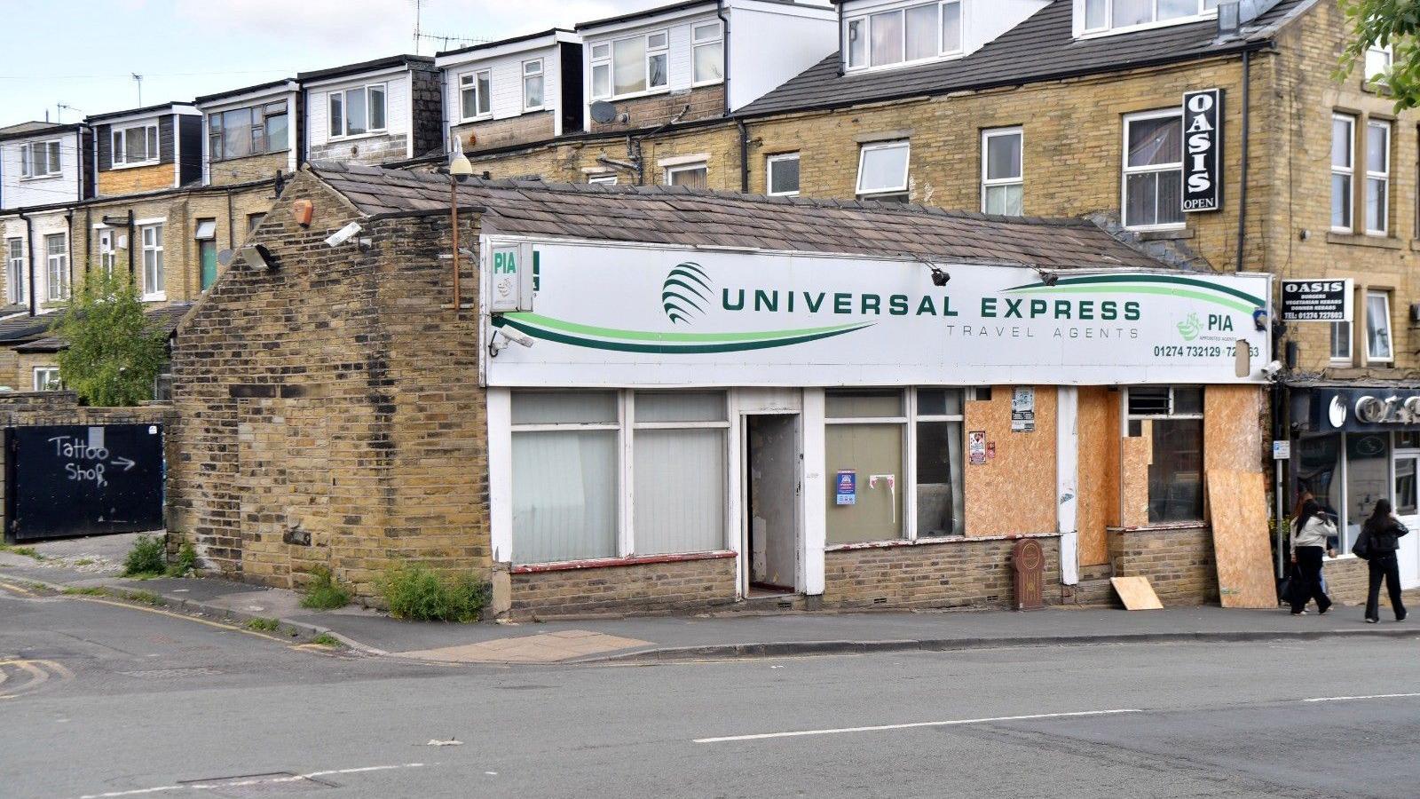 A single storey brick travel agents building. Some windows are boarded up and there is a memorial stone outside.