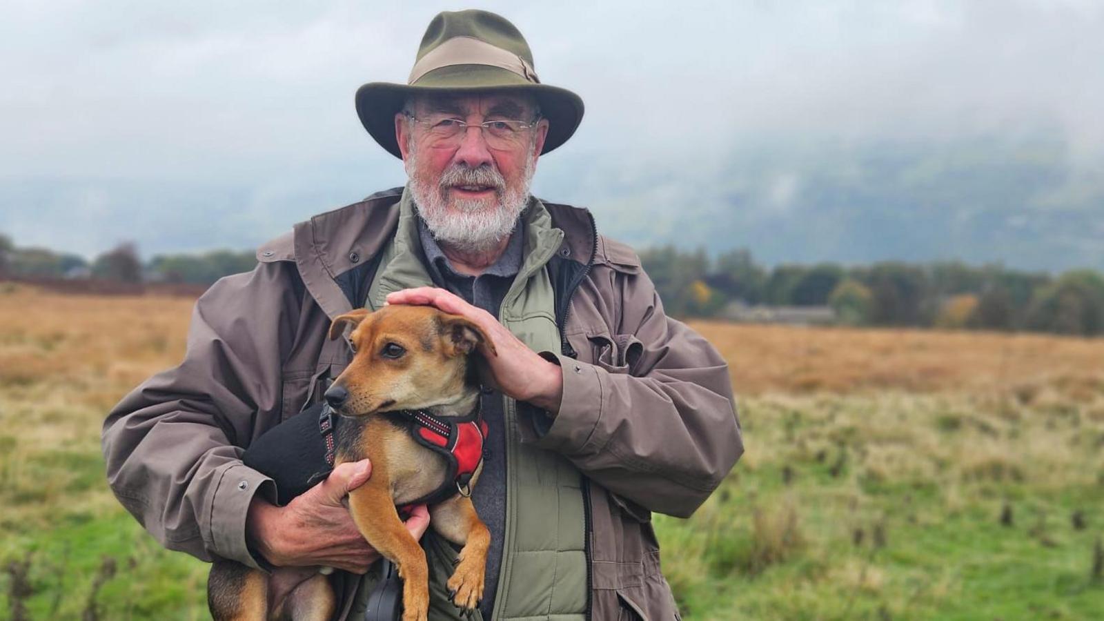 Owen Wells, from Friends of Ilkley Moor, with pet dog Bran. Owen is standing on a spot at Ilkley Moor, which can be seen in the background. He is wearing a green hat and outdoor clothing and is holding the dog.