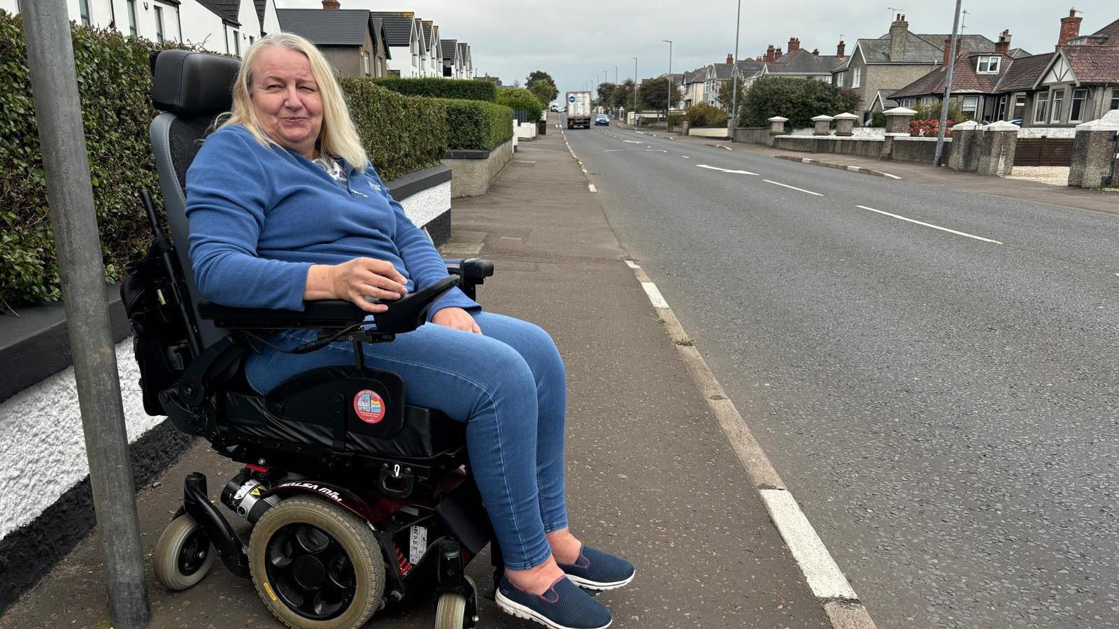 A woman in a motorised wheelchair looks off to the side of the camera. She has light blonde hair and is wearing a blue fleece top and blue jeans. She is at a bus stop on the side of a residential street. In the background you can see grey paving, homes and light traffic in the distance. 