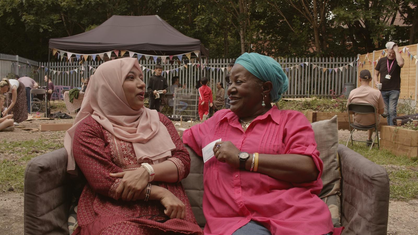 Who women sat on an outdoor sofa in a community garden. The woman on the left is wearing a pale pink hijab with red traditional clothing. The woman on the right is wearing a teal headwrap, and a bright pink shirt. They are facing each other and appear to be engaged in conversation