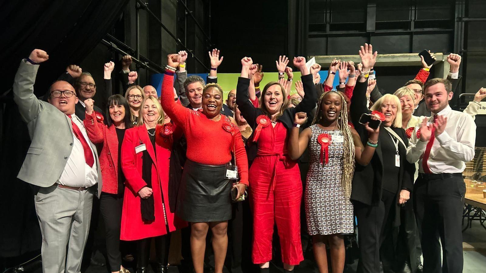 A group of people wearing red Labour rosettes, smile and put their hands in the air as they celebrate a win. 