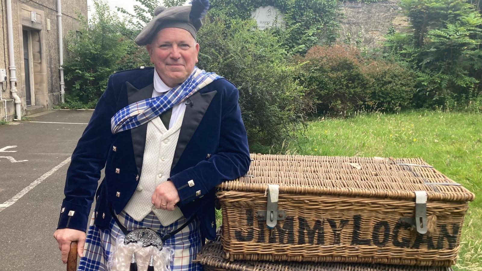 A man in a flamboyant, bright tartan dress outfit is leaning on a stick and against a large basket with Jimmy Logan written on the front. He is in a garden and he looks happy.