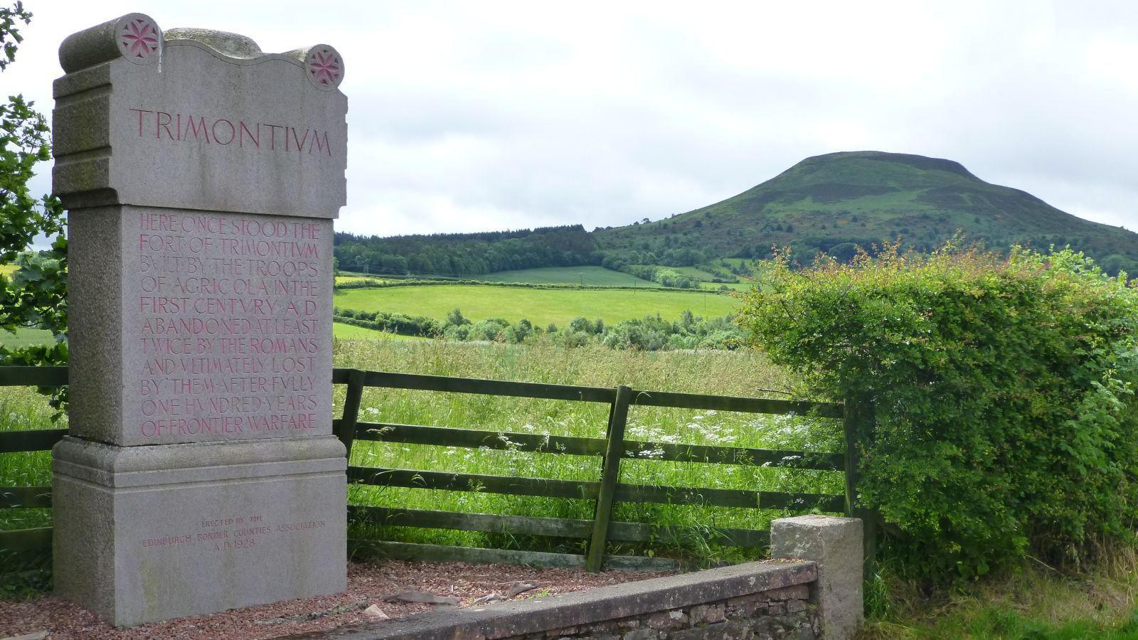 A large stone monument marking a site of the Roman occupation of southern Scotland with fields and a hill in the background