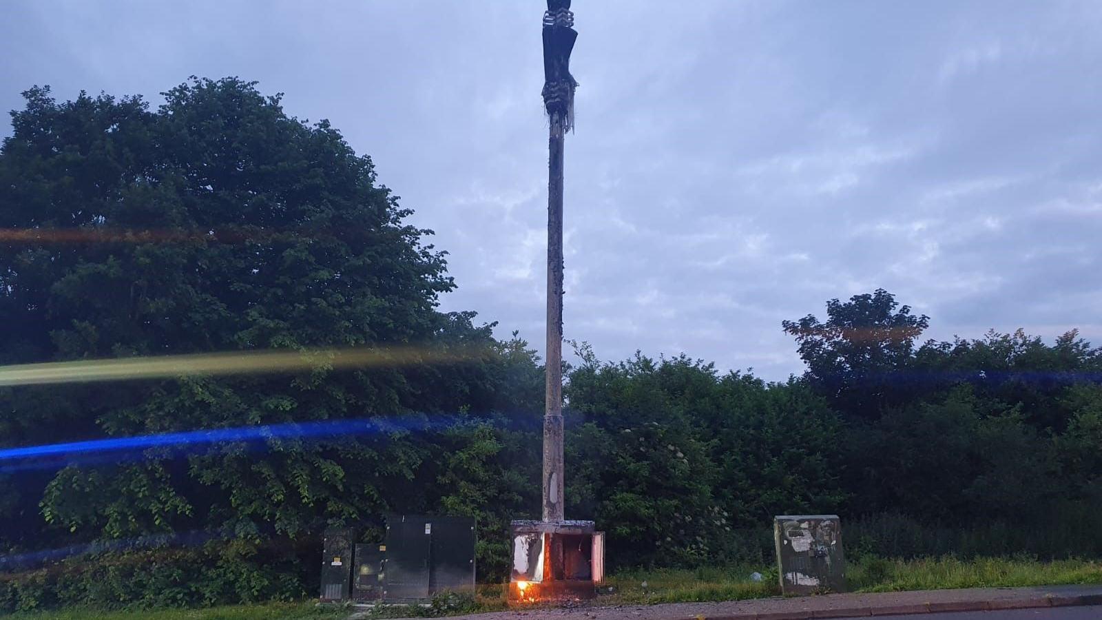 A 5G mast is on fire. Trees and bushed are in the background. It's late evening.