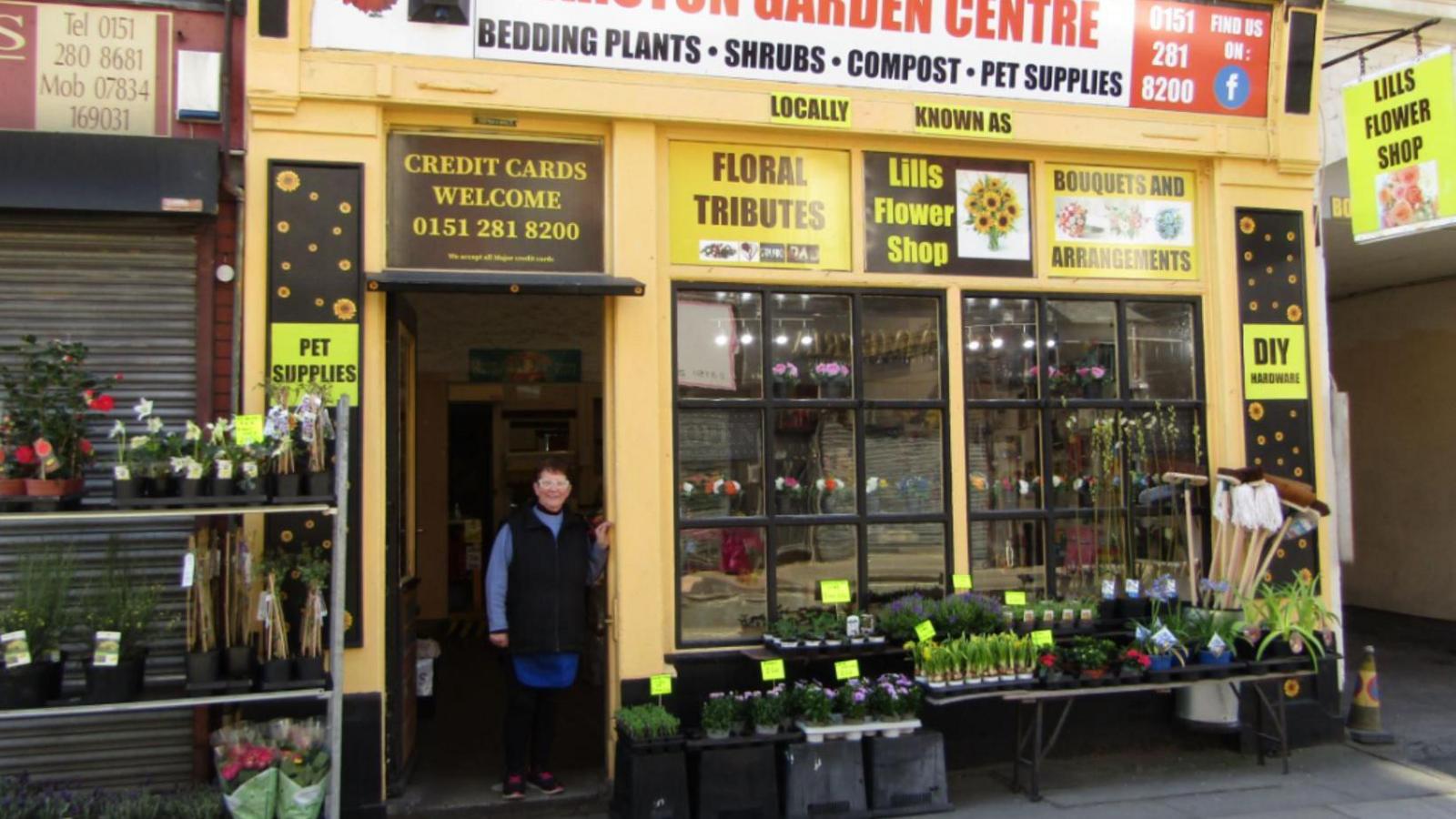 Garston Garden Centre shop front is painted yellow with flowers and plants outside. Lill is stood in the doorway smiling