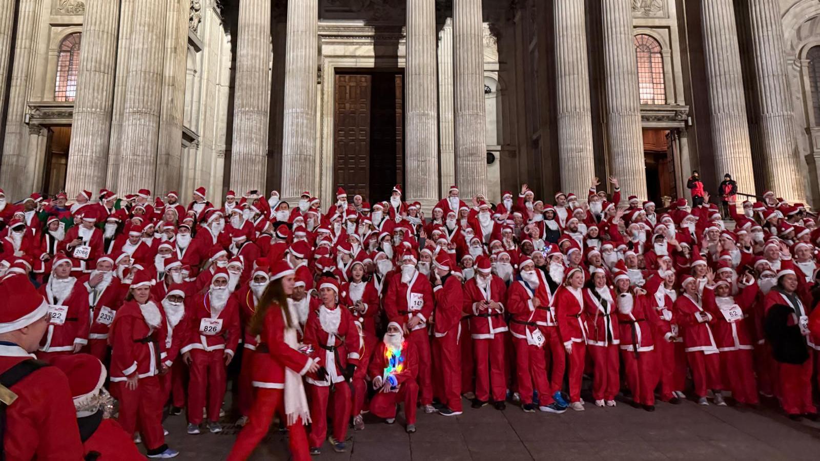 A large group of people dressed as Father Christmas cheering and waiting on some steps outside a large building with pillars. 