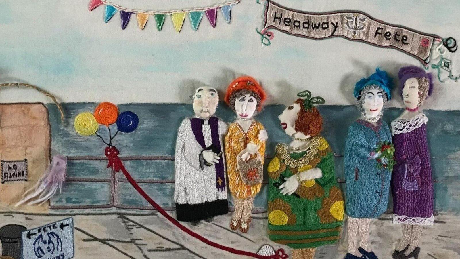 A close-up of some of the embroidery which is on display in Jersey. It shows three embroidered balloons which are yellow, orange and blue tied onto railings on a promenade in front of the sea. Next to them are five figures with some wearing dresses and hats. Above them is a sign that says Headway Fete.