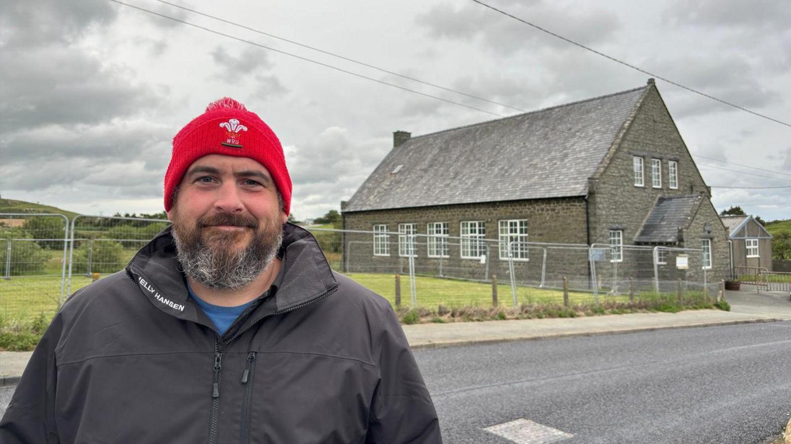 Huw Owen yn sefyll o flaen neuadd Mynytho