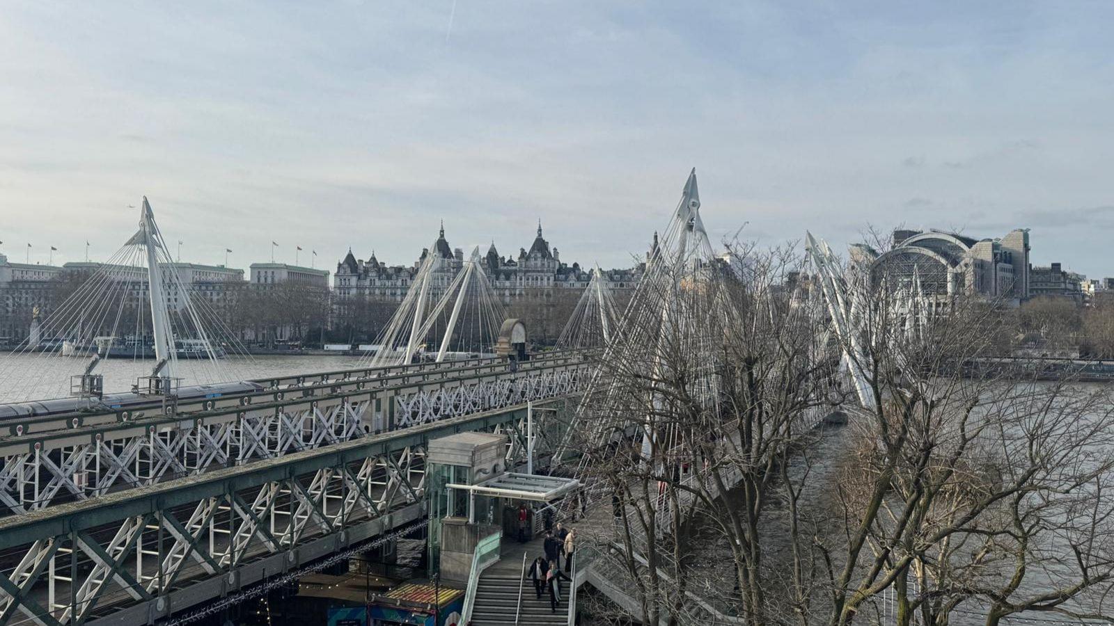 A landscape shot of the Victorian-style bridge over the River Thames, with a number of outward leaning pylons along each side. There is a pedestrian bridge beneath it.  Charing Cross is in the background, and tree branches are in the foreground of the picture.  