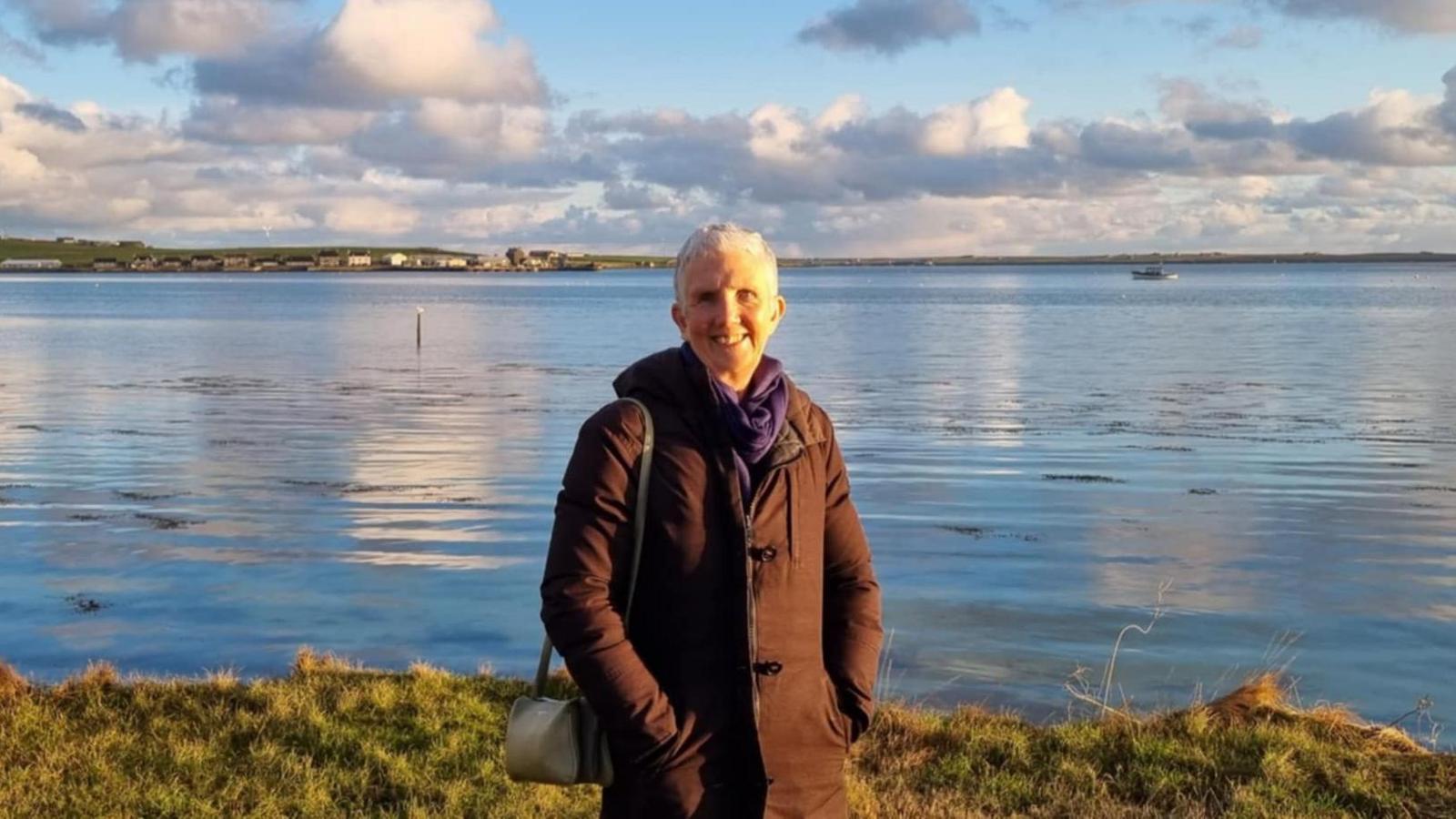 Ann Cleeves, wearing a brown jacket and purple scarf. She is smiling and standing in front of a body of water.