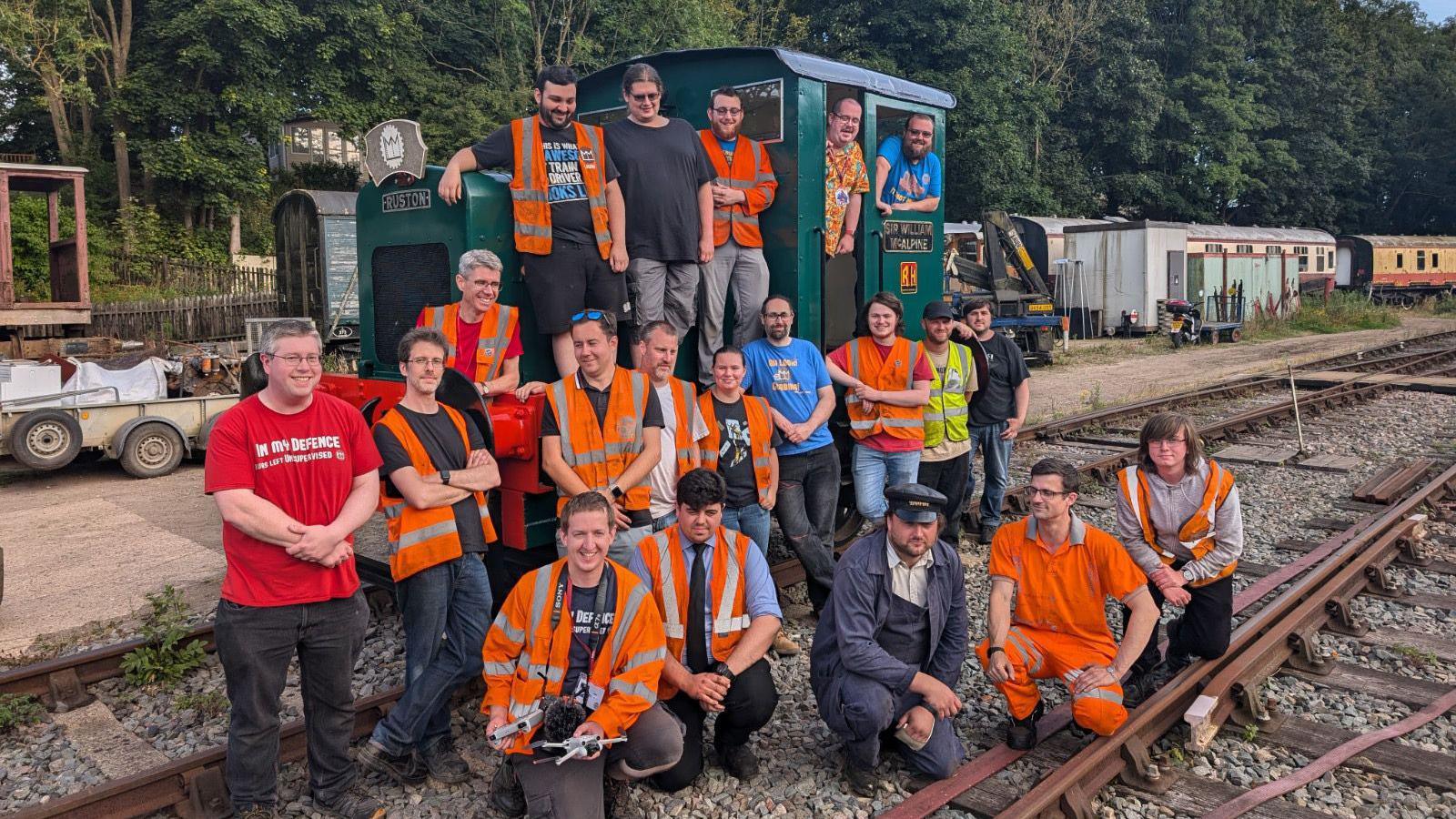 A large group of men stand around a green locomotive on train tracks. They are wearing orange hi-vis jackets and smiling at the camera. 