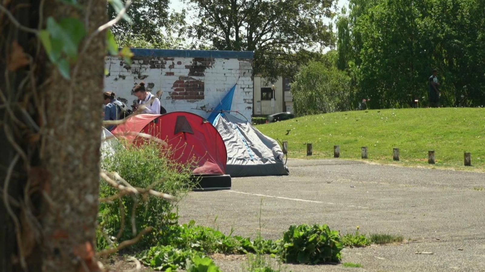 A grey and red tent in a park with homeless outreach workers in the background speaking to asylum seekers to support them