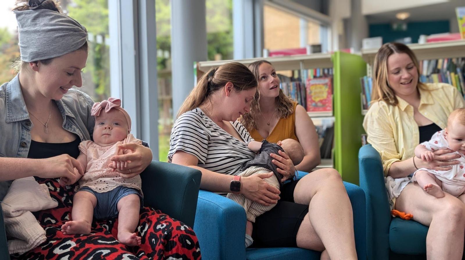 A group of four mums sat in blue armchairs with their babies in a room. There two rows of book shelves in the background and a large glass window.