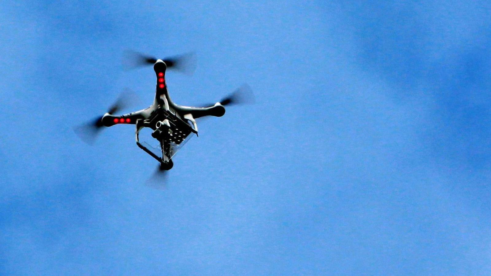 A drone flying in a blue sky. It is black, has four propellers and red lights on it.