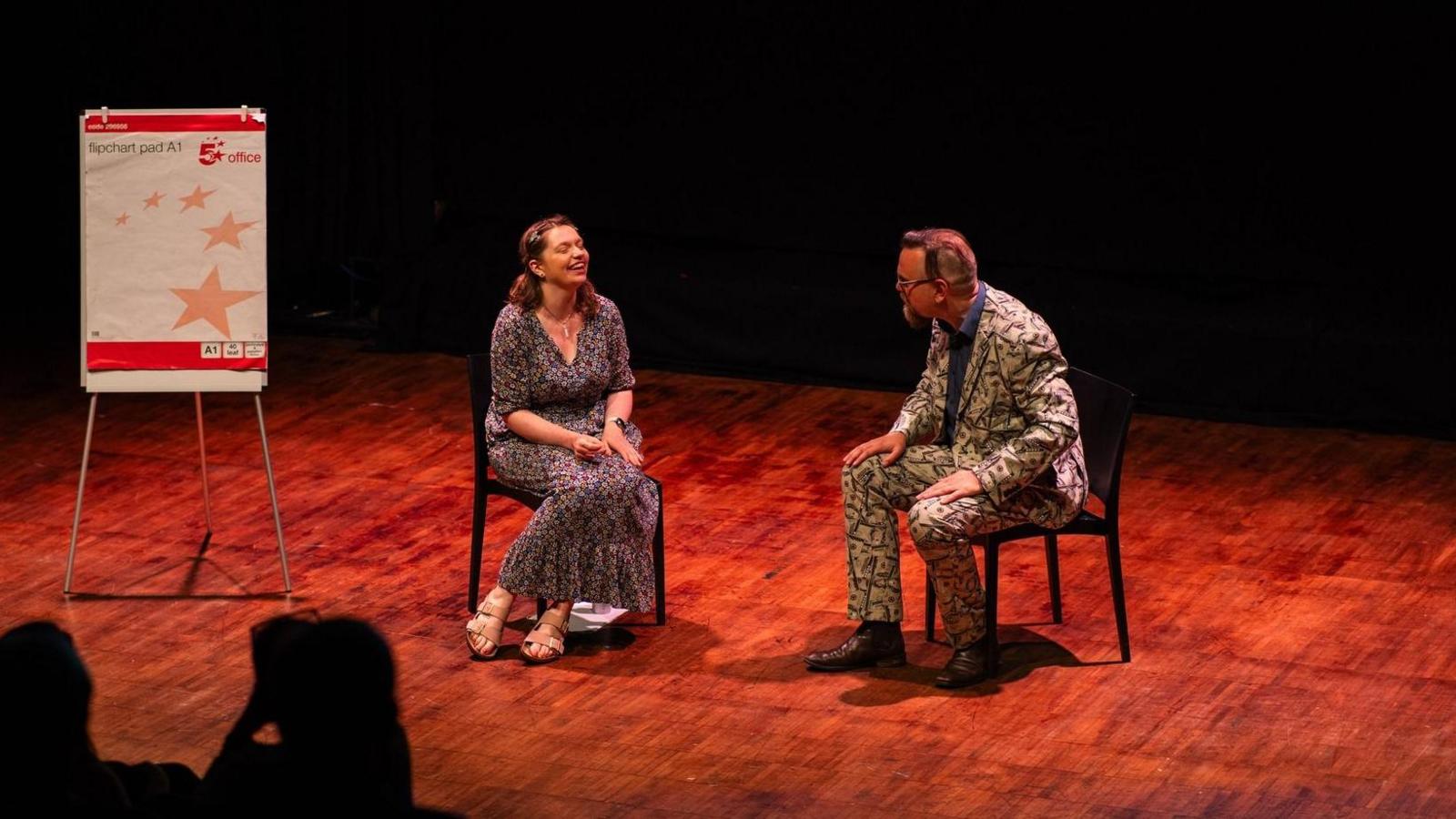 Isobel Jeffery sat in a chair on stage with another person in a chair opposite. Isobel is wearing a long dress and the other person a suit.