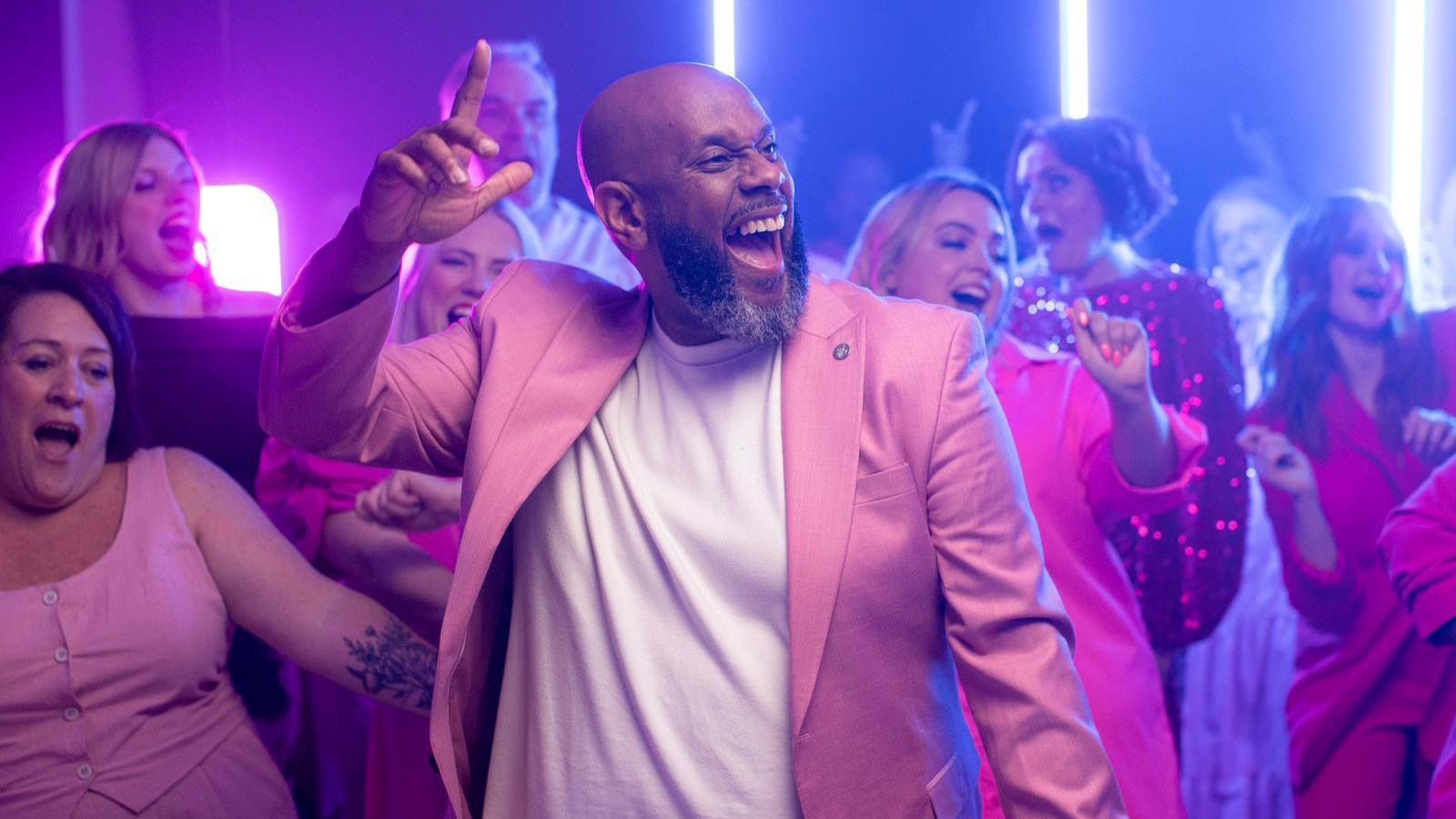 Gareth Fuller, in the middle, with his right arm help up, mid song, he is wearing a white T-shirt and pink jacket. He has a bald