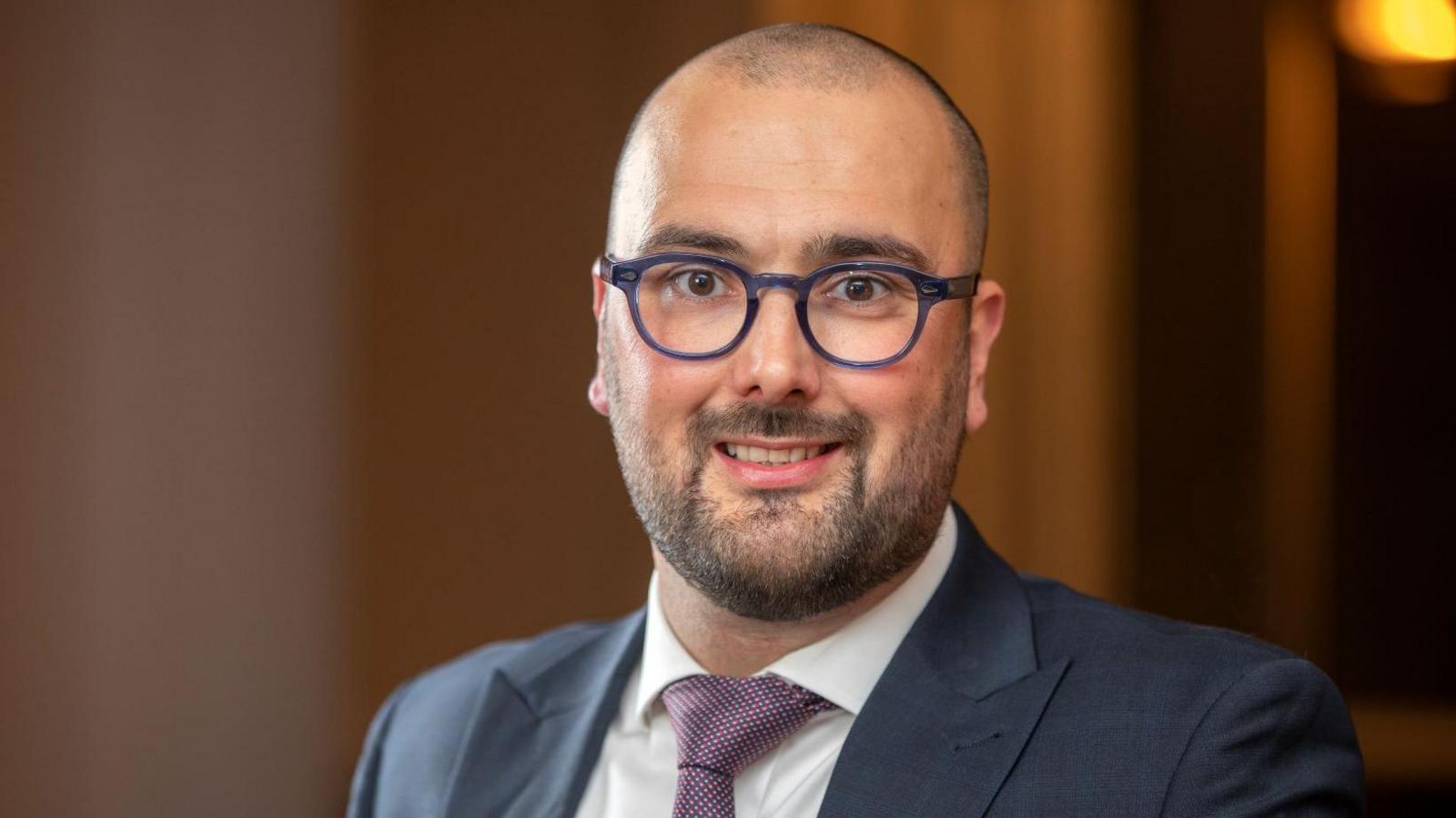 Photo of a man wearing a suit with purple tie. He has a bald head and is wearing purple glasses with stubble.