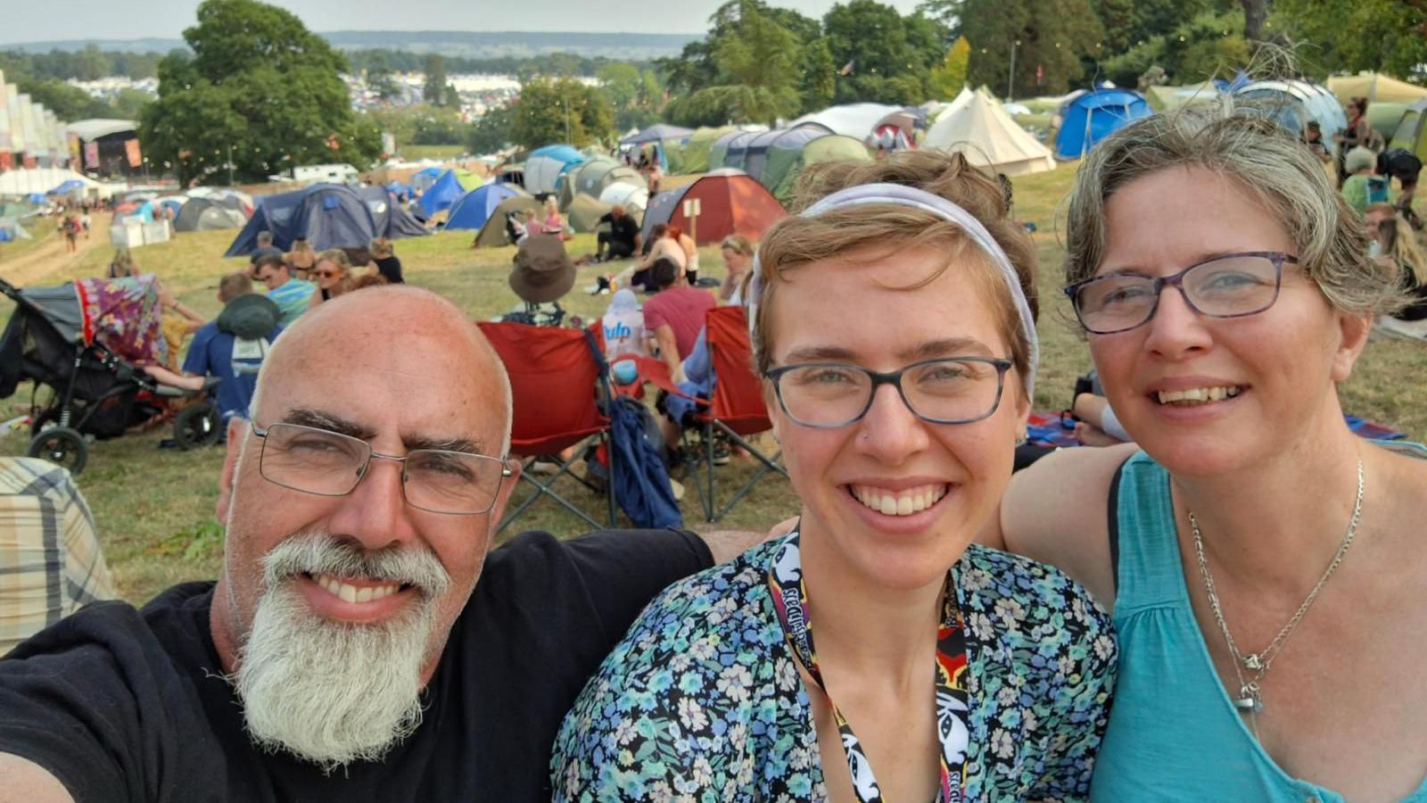Freya and her parents pose at a music festival in front of rows of tents. Freya is in the centre and is wearing a floral dress and a lanyard. She has glasses and short blonde hair pushed back with a headband. To her right is her dad, who is bald with a grey beard and black T-shirt. Her mum is to her left and has short hair like Freya, glasses and a turquoise T-shirt. All three are smiling into the camera.