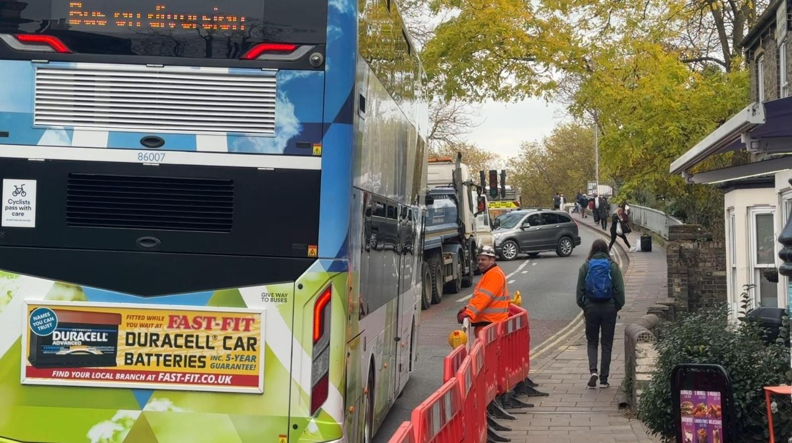 A double-decker bus to the left with a workman removing a road barrier to let it pass. Ahead is a car turning around. There are a number of trees with yellowy green leave above a road bridge. 