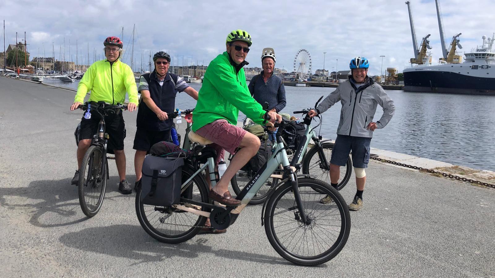 A group of five men with electric bikes, four in a row and one man front an centre sideways on, smiling at the camera. They are stationed in a harbour with the sea to the right.