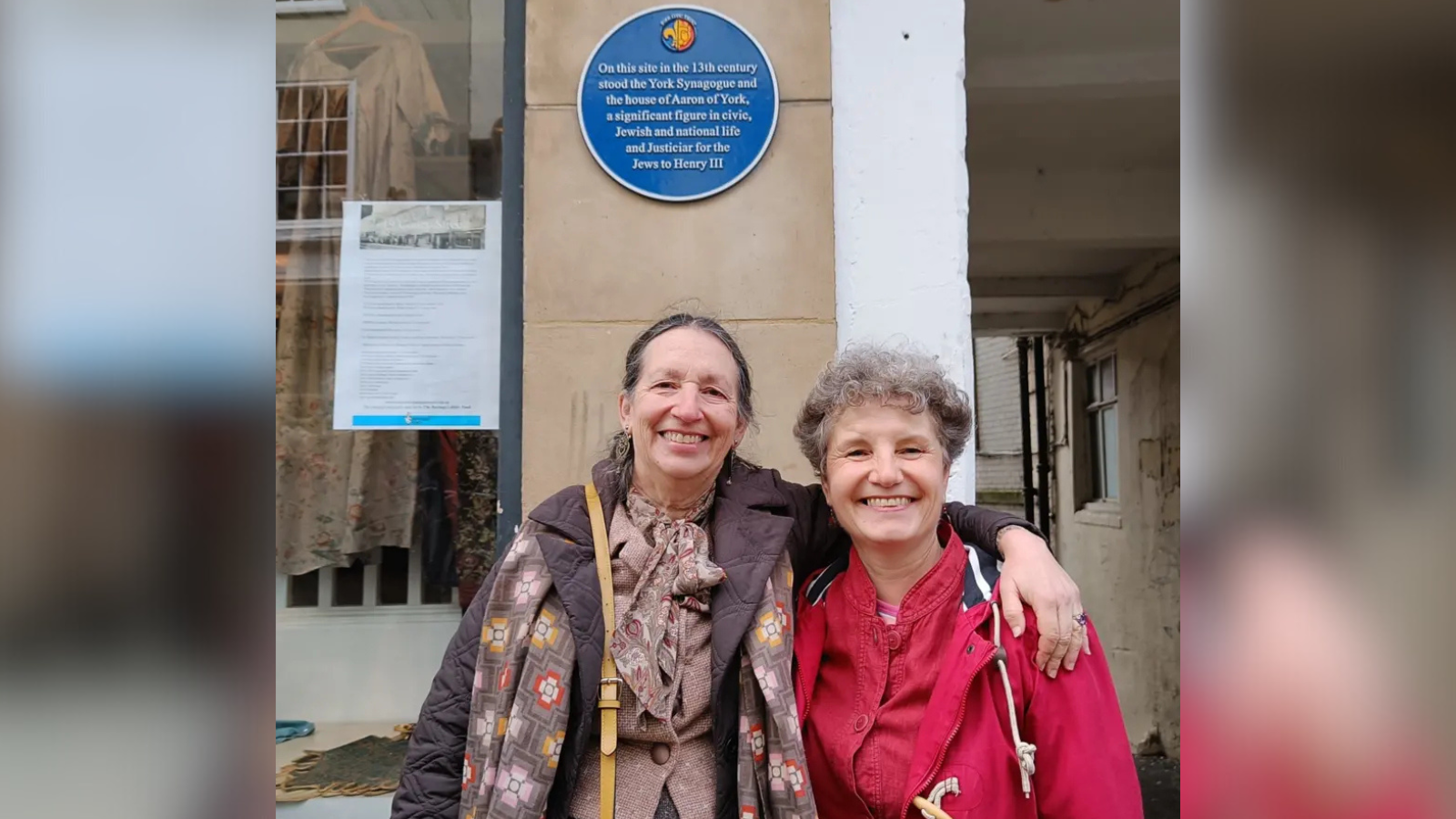 Rabbi Dr Elisheva Salamo (left) and Dr Louise Hampson (right) stand in front of the blue plaque on Coney Street in York. Rabbi Dr Elisheva Salamo wears a brown coat and a light pink jacket, and Dr Louise Hampson wears a red coat.
