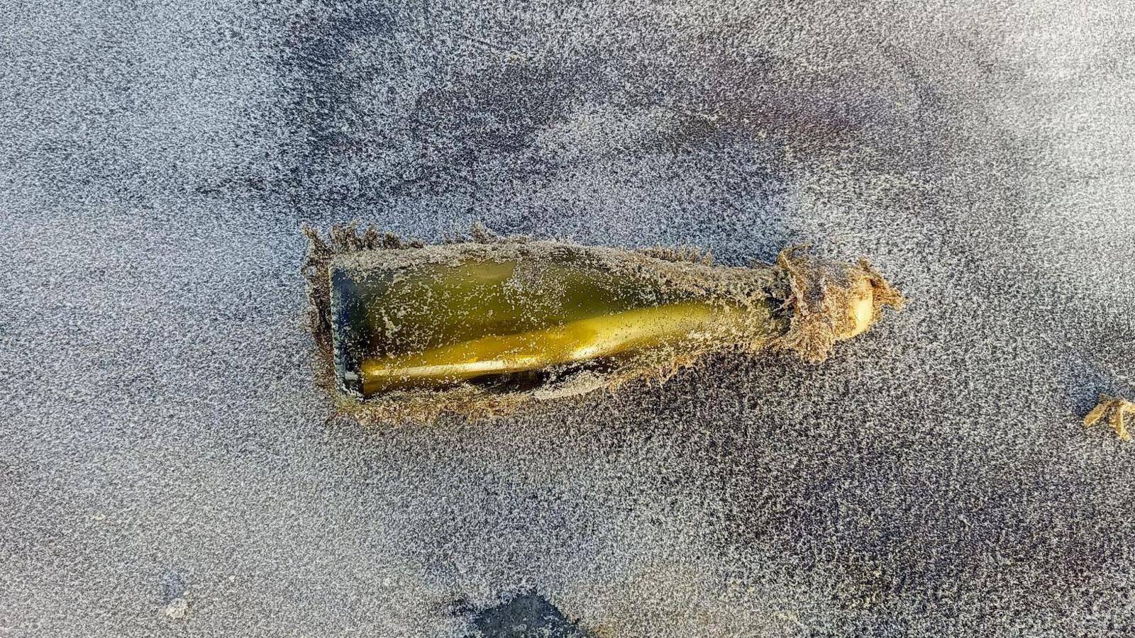 Blake's bottle lying on a beach. It is a green glass bottle with a white stopper. It is covered in sand.