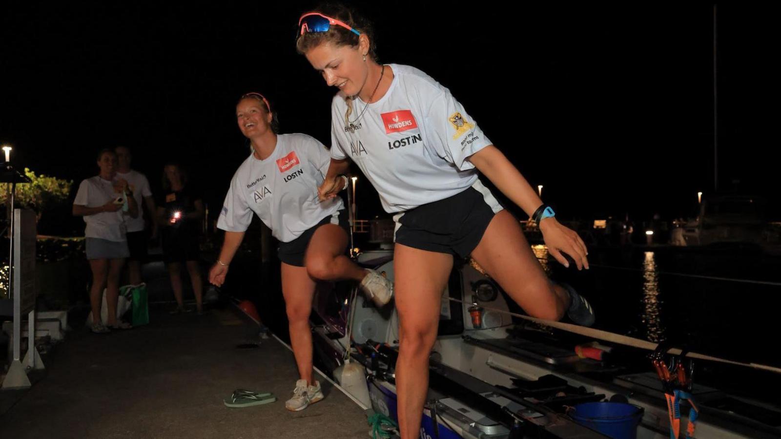 Two women step off a small rowing boat on to the harbour side at Cairns, it is dark and a small crowd has gathered to welcome them. The women are wearing identical outfits of black shorts and white t shirts with sunglasses perched on their heads. They are both smiling and are holding hands.