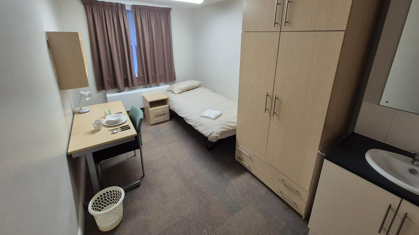 A room with a bed in it with cream covers. There is a desk to the left with crockery and a lamp on it. There is a wardrobe by the bed, and a sink with cupboards below it and a mirror above. There is a cream bin on the left and a green chair at the desk. There is also a bedside table.