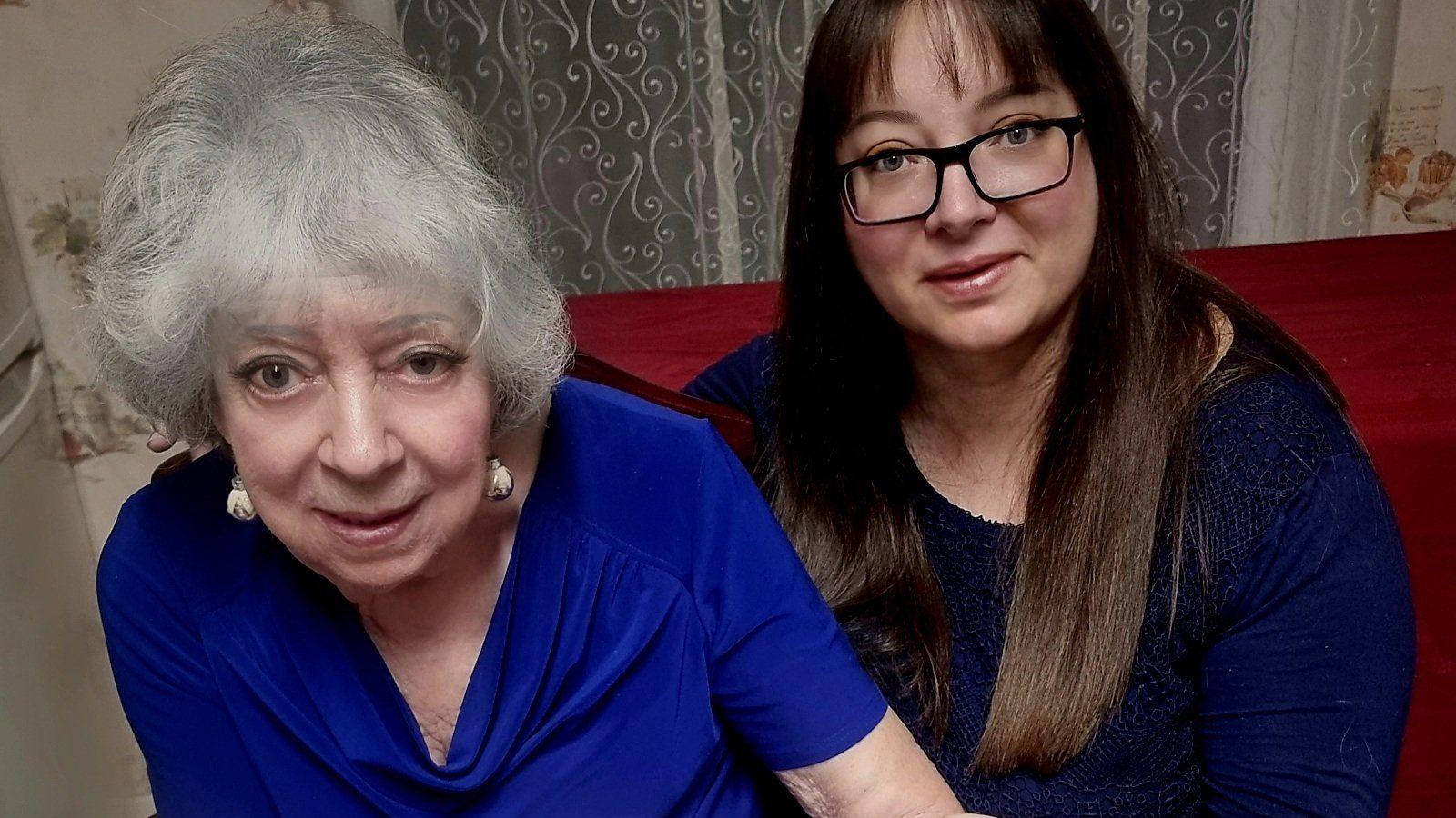 A woman with white hair and cobalt-blue top and a woman with brown hair in a dark blue jumper sitting on a red sofa.