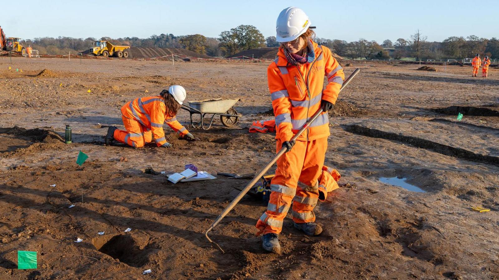 An archaeologist wearing fluorescent orange overalls and a white hat is holding a hoe and digging in the bare soil as another archaeologist, kneeling down, is working beside a wheelbarrow 