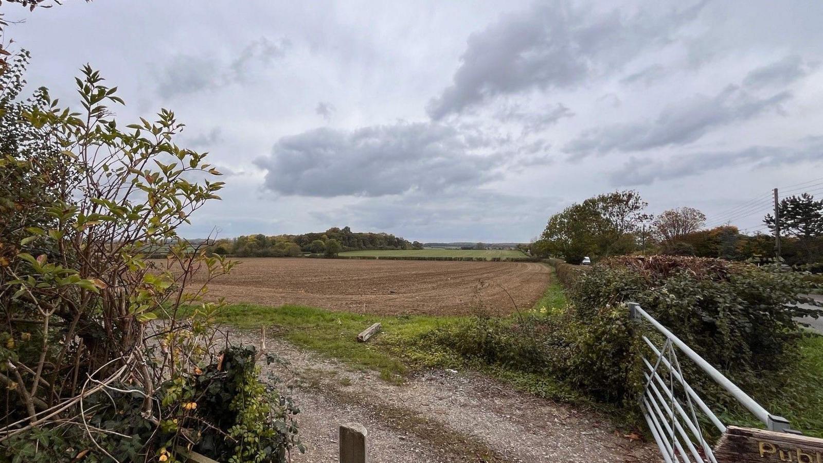A large brown ploughed field surrounded by hedges, seen from a metal fence at the corner.