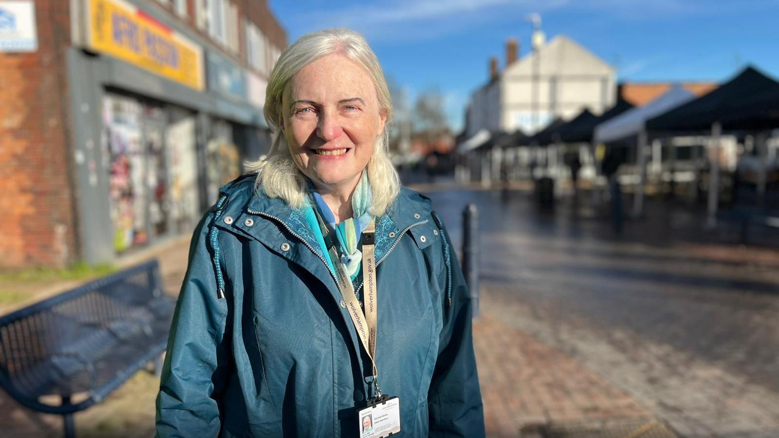 A woman with white, blonde hair to her shoulders smiles at the camera while stood on a high street. She wears a blue coat with a beige lanyard around her neck, holding some sort of ID card. The street has an iron bench, temporary marquees for a market and buildings with shops in behind her.