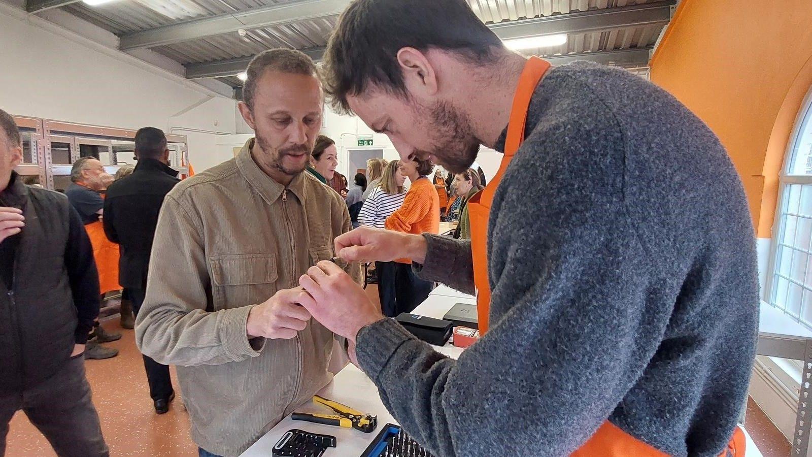 In the foreground, two men fix a small device inside a warehouse. One of them is wearing a beige shirt, the other an orange apron over a blue jumper. A tool is on the desk between them and people are milling around in the background