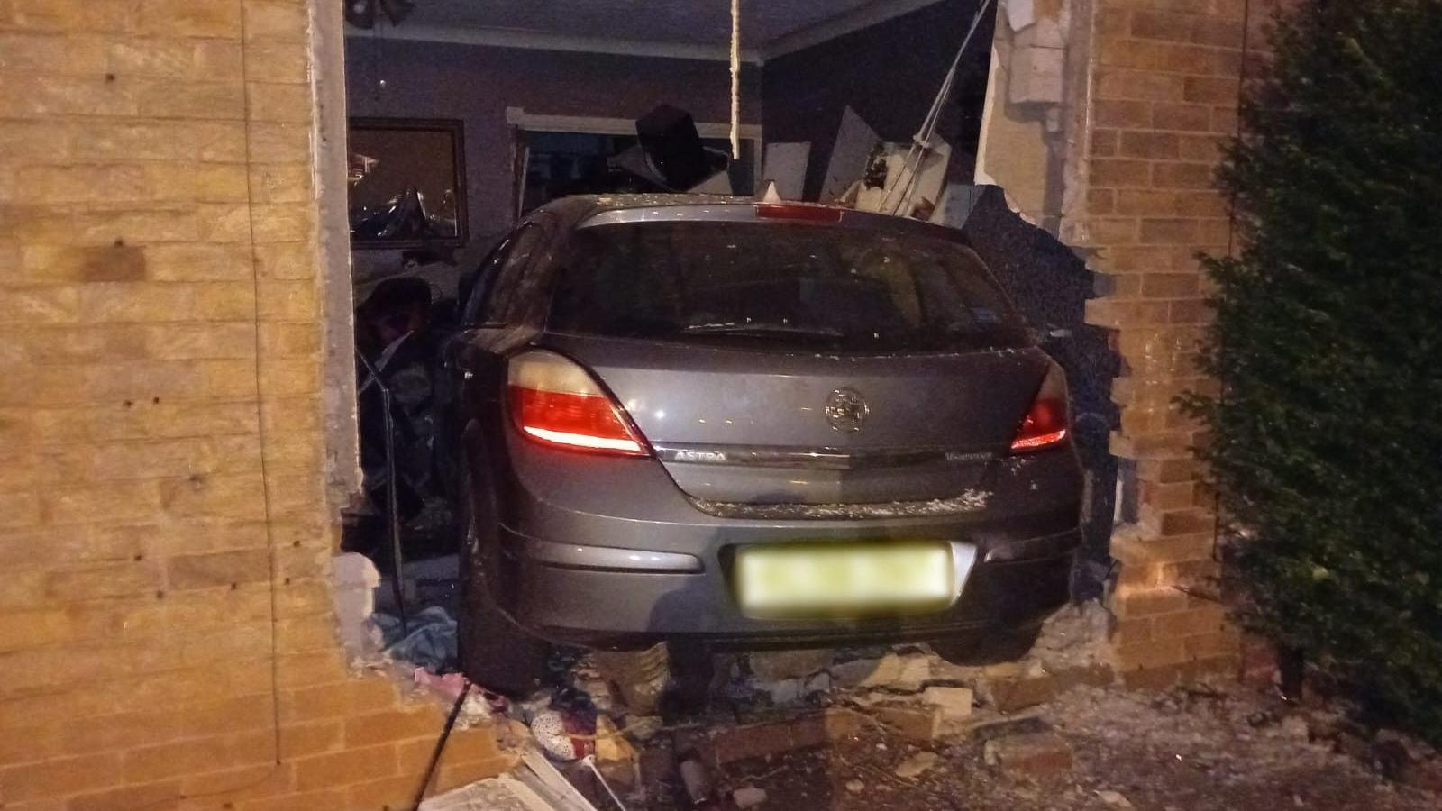 A grey Vauxhall Astra that has crashed into the wall of a house leading to a living room. Bricks and debris surround the car