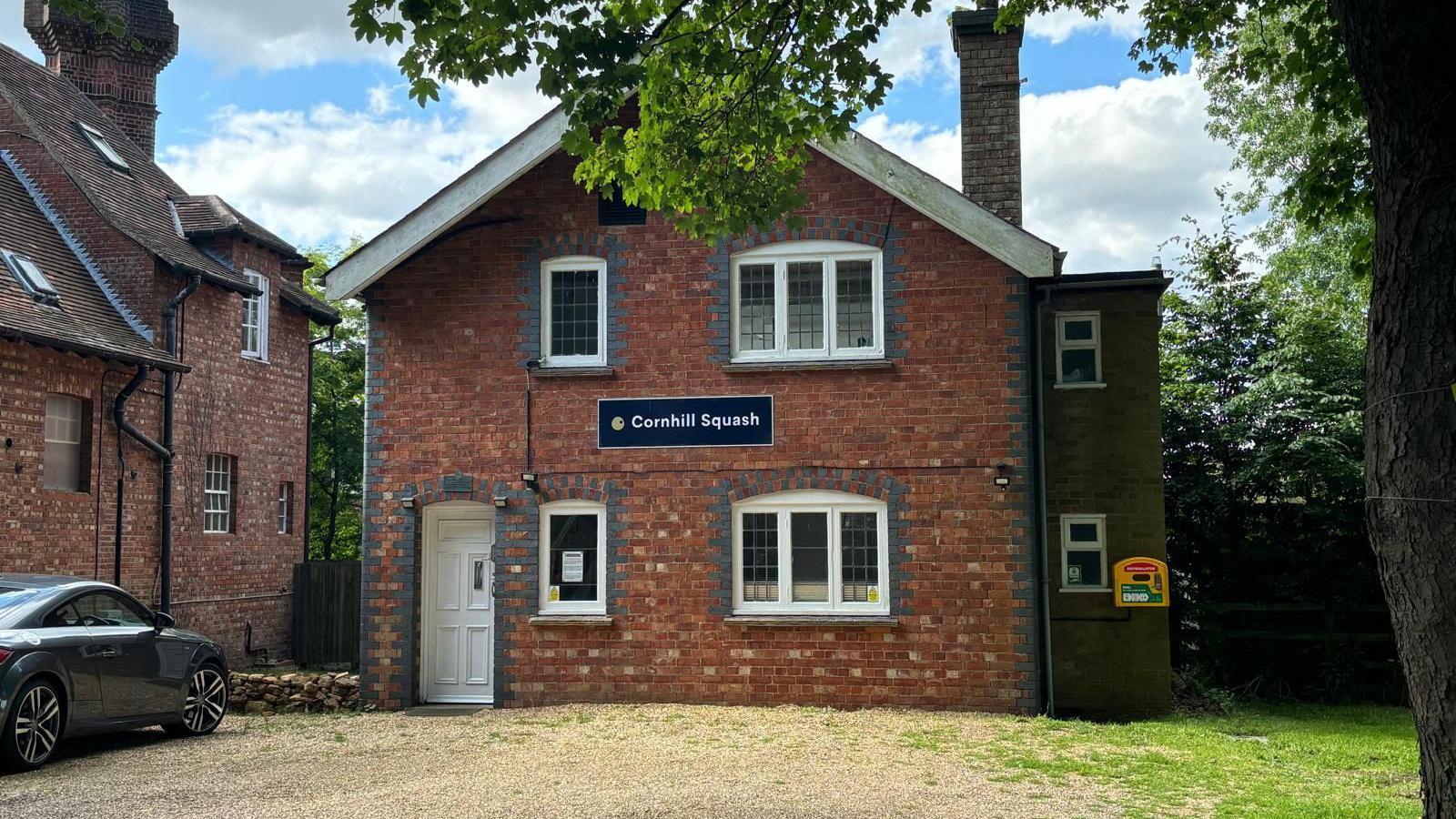 The outside of Cornhill Squash club -  a red brick building with a white door and a navy sign with "Cornhill Squash" written on it. 