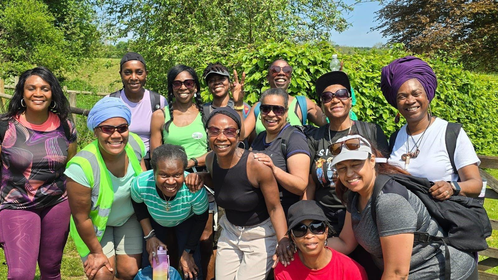 A group of 13 women of colour posing for a group photo on a sunny day. They are standing in front of a wooden gate in a field, with bright green hedges and trees behind them. They are wearing brightly-coloured walking clothes and many have their arms around each other.