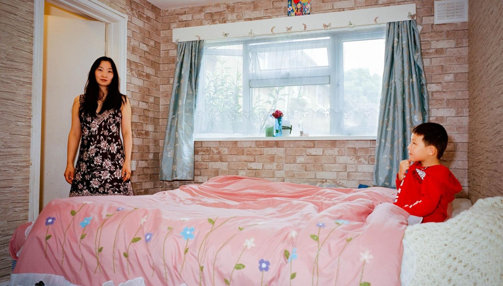 A woman stands on the left and a child sits in bed on the right. The room has neutral pink tones in it and a window is seen between the two people.