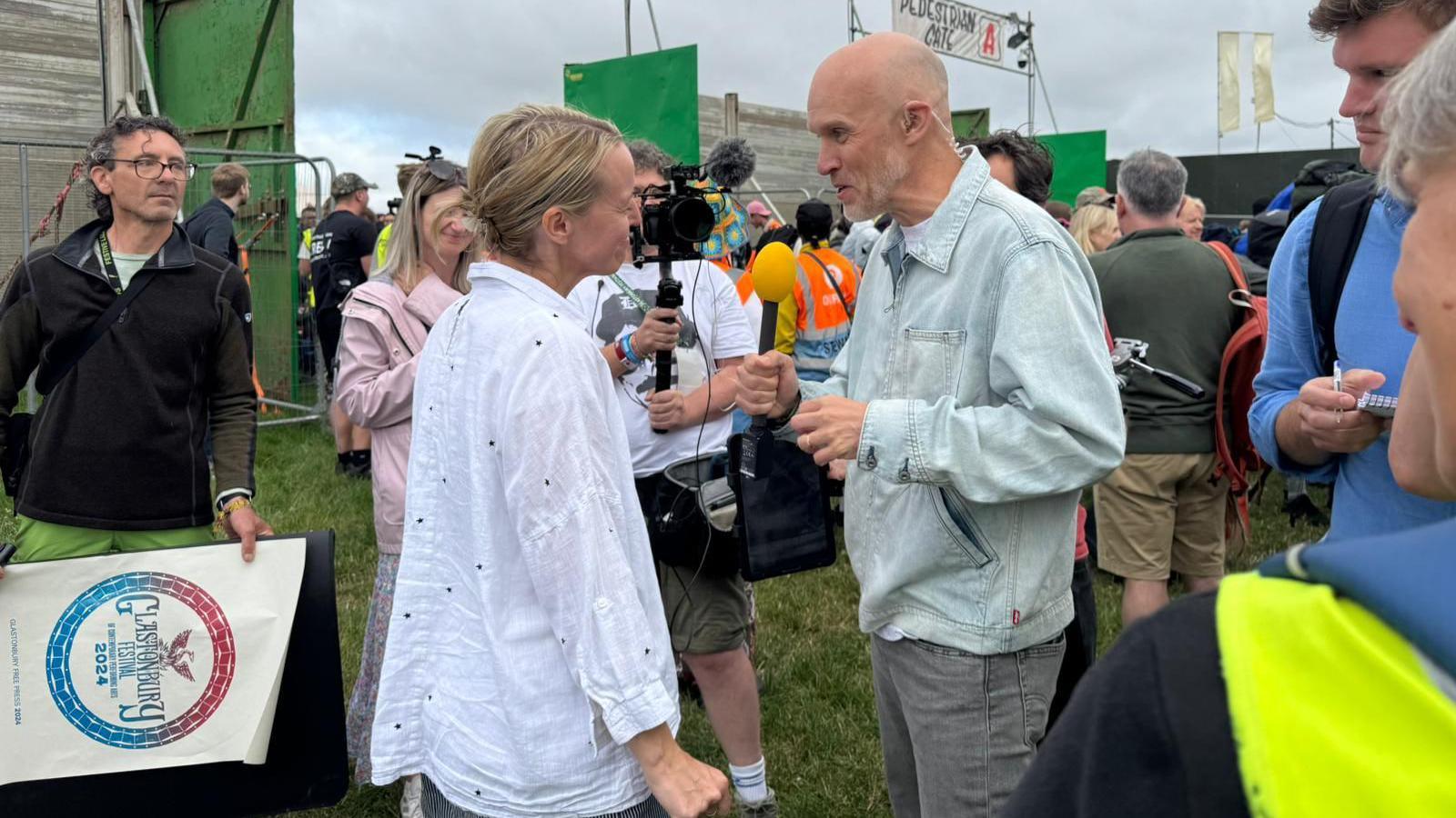 Emily, shown wearing a white shirt and with her blonde hair back, being interviewed by a BBC News journalist. There are lots of other journalists surrounding her. Crowds can be seen in the background as they enter the gates to Worthy Farm.