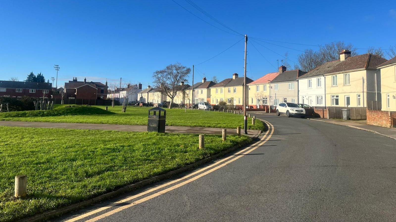 A road bend next to a children's park with a footpath running between it. On the other side of the road there's a row of attached houses, a couple of them have white cars parked in front of them. It's a sunny bright day with a clear blue sky.