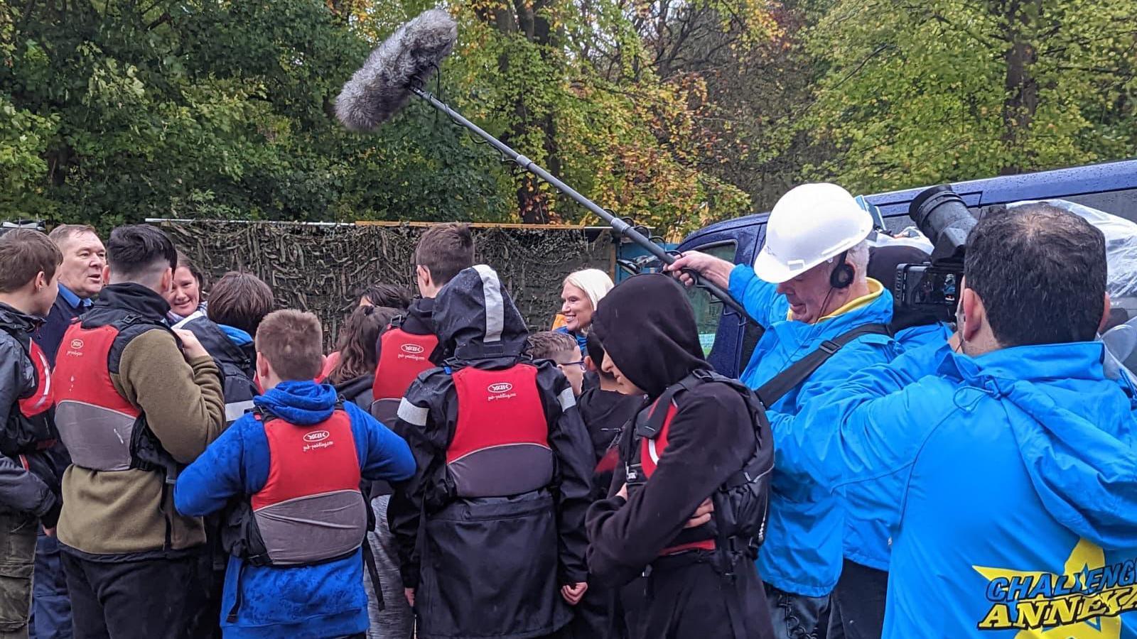 A group of young cadets in red life jackets huddled and looking away from the camera. Anneka's face and blonde hair can be seen through the crowd. In the foreground are two men in blue waterproof jackets who are filming the scene.