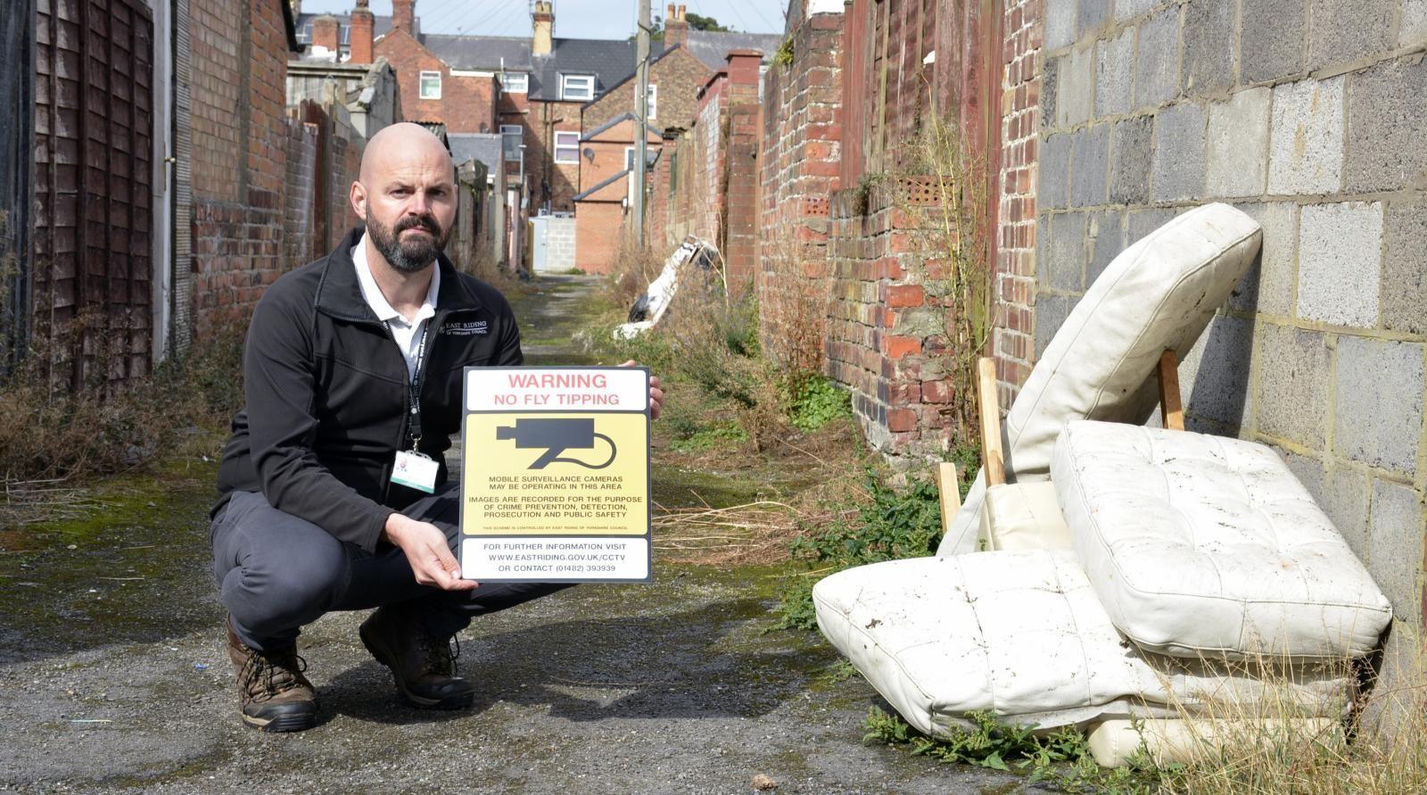 A council officer wearing a black fleece and grey trousers crouches in an alleyway next to a discarded chair and white cushions. He is carrying a sign warning mobile surveillance cameras are in operation.